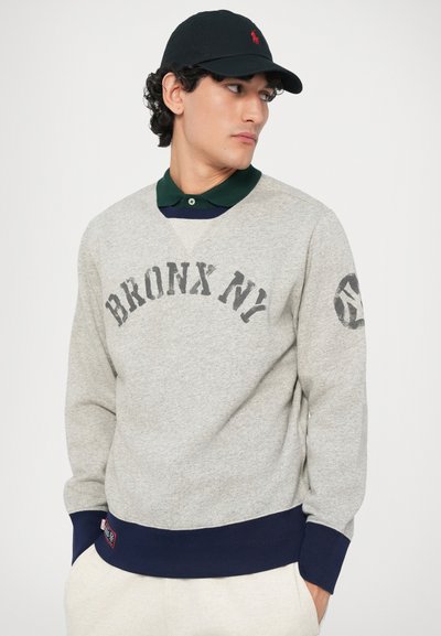 Mladenič nosi sivo majico s napisom "BRONX NY", temno zeleno srajco s ovratnikom, črno kapo z rdečim logotipom in stoji z rokami v žepih.