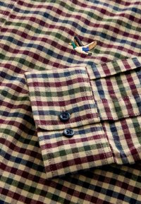 Karierte Hemdmanschette mit blauen und dunklen Knöpfen, verziert mit einem gestickten fliegenden Stockenten-Motiv auf einem Stoff mit grünen, blauen und burgunderfarbenen Karos.