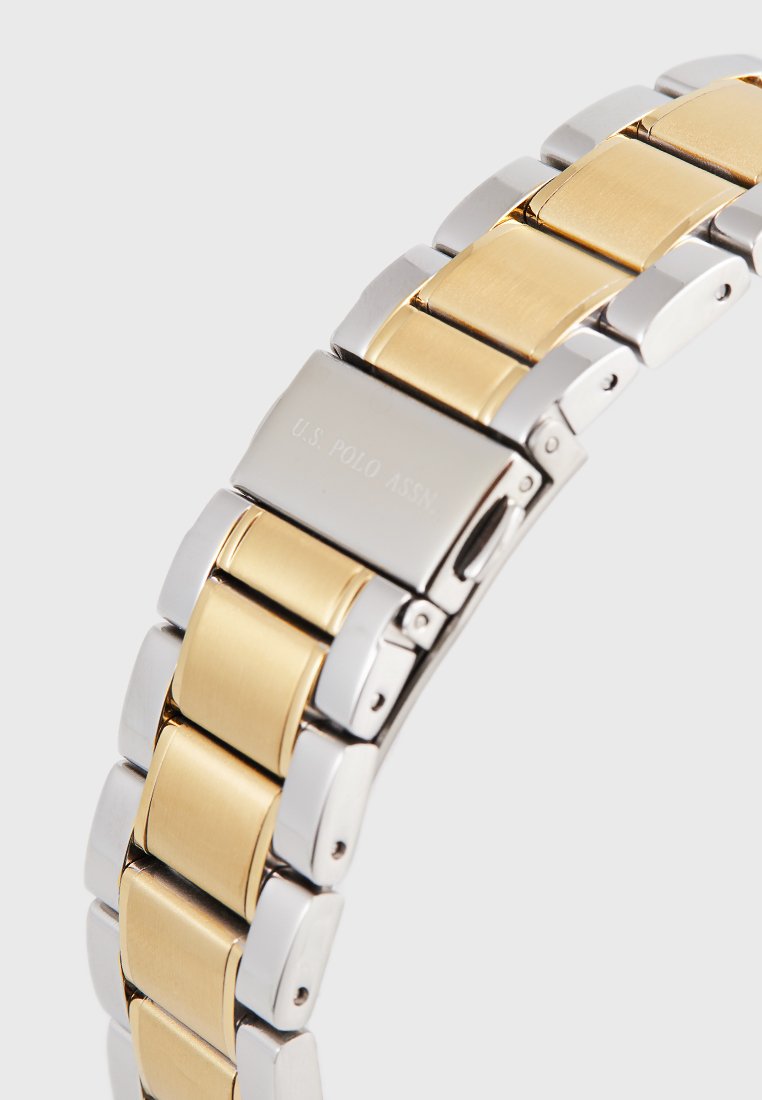 Pulsera de reloj de metal en plata y oro con eslabones rectangulares, que cuenta con un acabado pulido y un cierre grabado con "U.S. POLO ASSN."