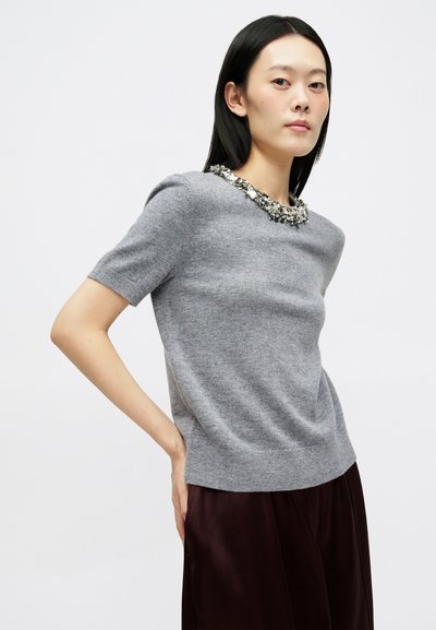 Siva pulover iz mehkega materiala, s kratkimi rokavi in dekorativnim okrasom na izrezu. Združen z temnimi satenskimi hlačami.