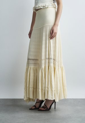 Femme portant une jupe maxi en dentelle crème à volants et des sandales noires à talons hauts et à lanières, sur un fond uni.