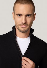 Czarny sweter na zamek z wysokim kołnierzem, o prążkowanej strukturze i brązowymi detalami zamka, noszony na białej koszulce.