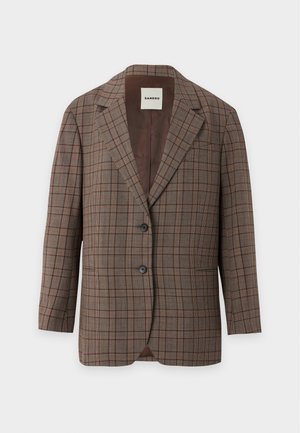 Kvinde stående iført en brun ternet blazer, cremefarvet top, beige vide bukser med små prikker, sorte sko og holder en lille brun håndtaske.