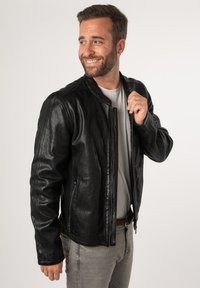 Giubbotto di pelle nera con una finitura leggermente texturizzata, chiusura con zip e due tasche laterali. Indossato sopra una camicia grigia chiara e pantaloni grigi.