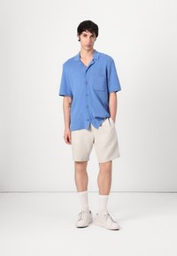 Camisa de botones de manga corta en azul con un bolsillo en el pecho, combinada con pantalones cortos beiges. El modelo lleva zapatillas blancas y calcetines cortos.