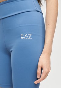 Γυναίκα που φοράει μπλε αθλητικά σορτς με ψηλή μέση και λευκό λογότυπο EA7 Emporio Armani στο αριστερό μηρό, στέκεται με το χέρι χαλαρό στο πλάι.
