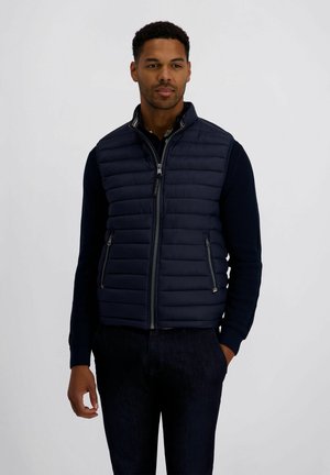 Mann trägt navyblaue gesteppte ärmellose Weste über dunklem langärmligen Hemd, steht mit einer Hand in der Tasche vor einfarbigem Hintergrund.