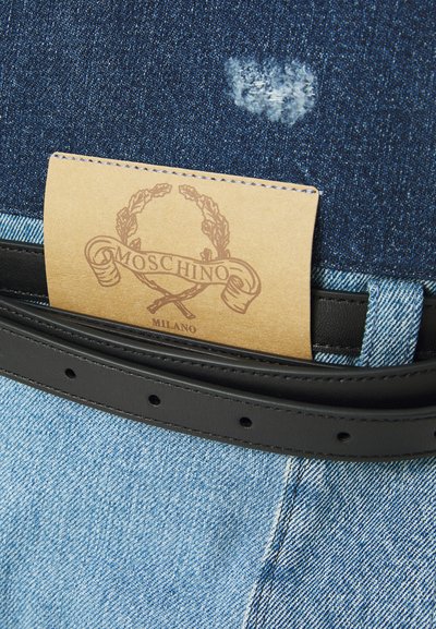 Eticheta maro Moschino Milano coaptată pe blugi negri de denim, deasupra unor blugi din denim deschis, cu un curea din piele neagră trecută prin bucle.