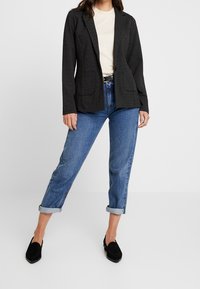 Svart strukturerad kavaj med två framfickor, kombinerad med en ljusbeige t-shirt och uppvikt blåjeans. Svarta loafers fulländar outfiten.