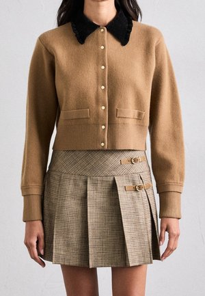 Femme portant un cardigan camel marron boutonné avec un col noir et une mini-jupe plissée beige à carreaux avec des détails de boucle.