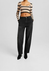 Kobieta ubrana w beżowy i czarny pasiasty krótki top oraz luźne czarne jeansy z wysokim stanem, w czarnych szpilkach, z rękami w kieszeniach.