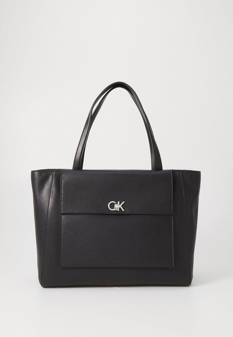 Sac cabas en cuir noir avec doubles anses et poche avant avec logo CK argenté sur un fond neutre.