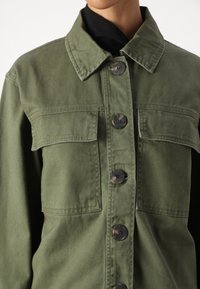 Zelená olivová bunda z bavlny s zapínáním na knoflíky, dvěma kapsami na hrudi a špičatým límcem. Strukturální a odolná tkanina.