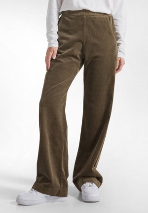 Persoon draagt bruine corduroy wijde broek met elastische taille en witte sneakers tegen een witte achtergrond.