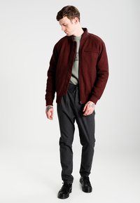 Giacca burgundy con zip e colletto alto, tessuto a trama, e tasca anteriore, abbinata a pantaloni jogger grigio scuro e sneakers nere.