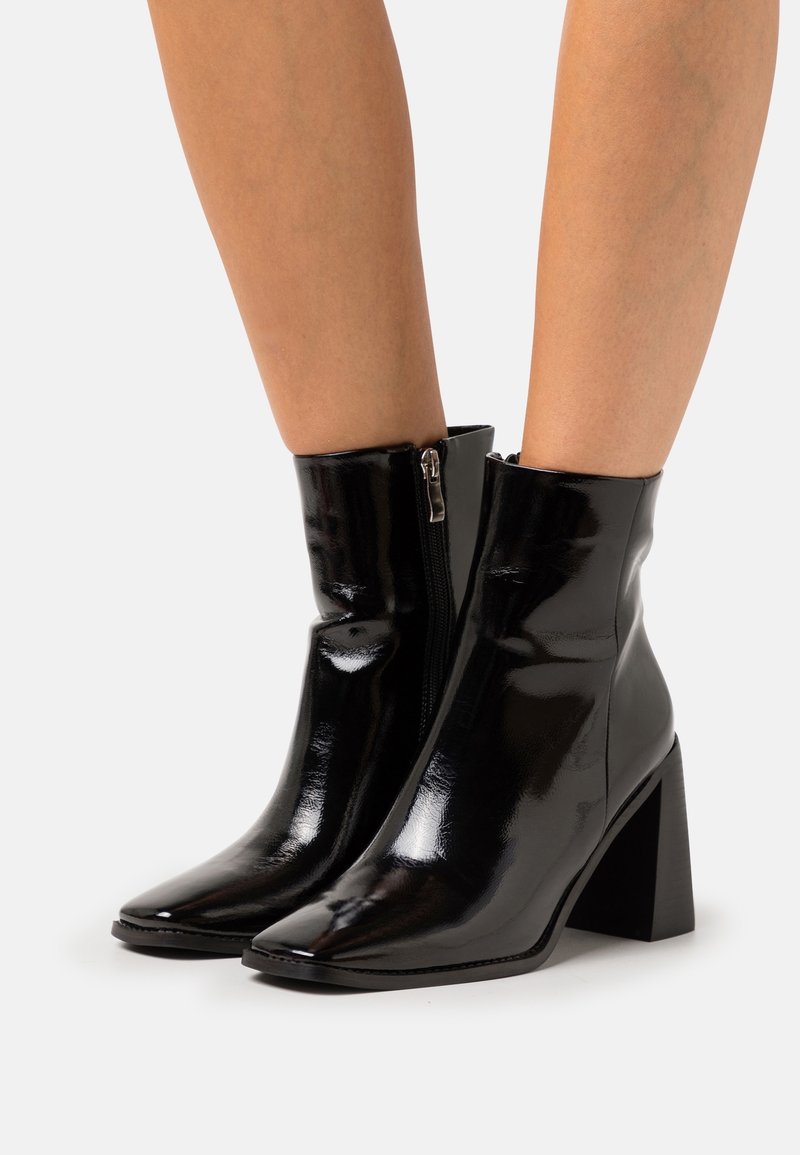 Bottines en cuir verni noir brillant à bouts pointus et talons blocs portées sur jambes nues sur un fond clair uni.