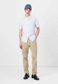 Camicia a maniche corte, blu e bianca a righe, pantaloni beige, scarpe scure e un cappellino azzurro chiaro. Il modello sta in piedi con una mano in tasca.