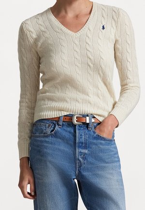 Personne portant un pull en maille torsadée couleur crème avec un petit logo bleu et un jean bleu avec une ceinture marron, une main dans la poche.
