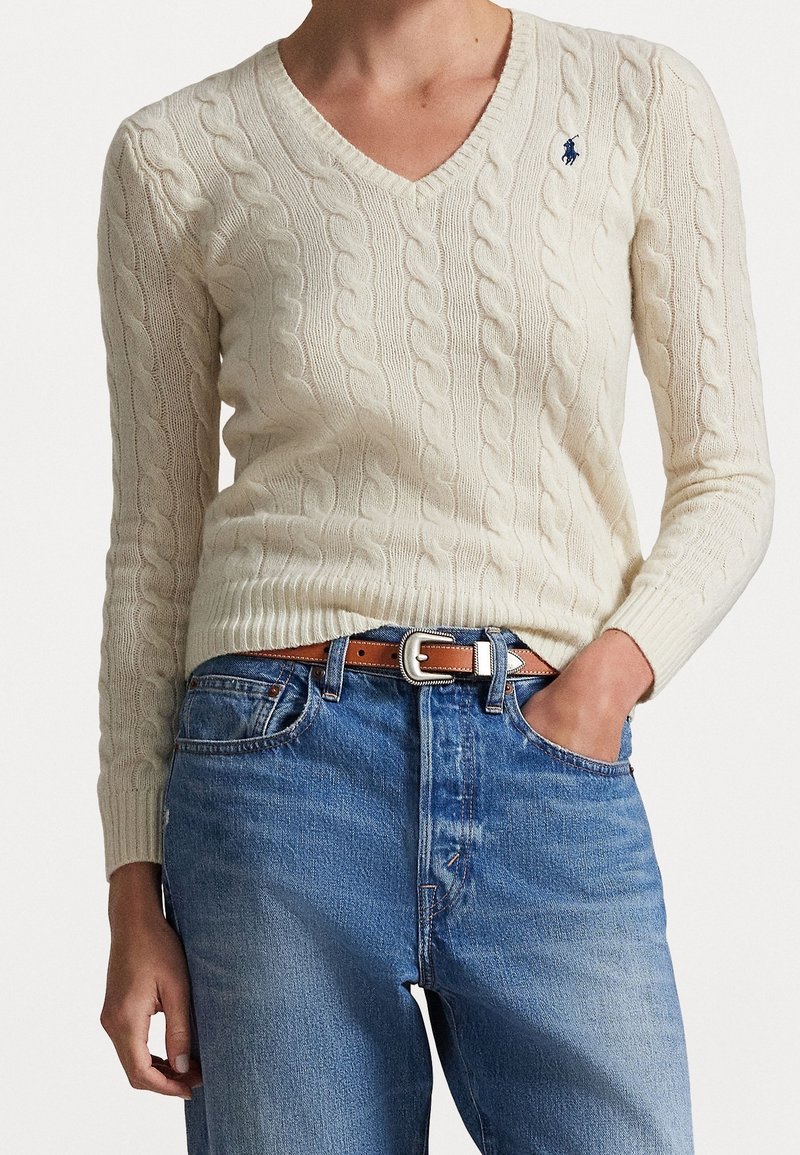 Personne portant un pull en maille torsadée couleur crème avec un petit logo bleu et un jean bleu avec une ceinture marron, une main dans la poche.