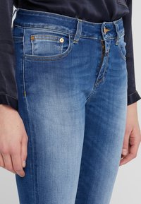 Jean bleu slim fit en denim. Caractéristiques d'un design à cinq poches avec un délavage clair, des accessoires dorés et des détails cousus le long des coutures.