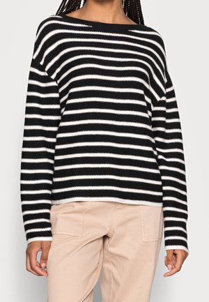 Schwarz-weiß gestreifter Pullover mit einem Bootsausschnitt und herunterfallenden Schultern, gestrickte Textur, kombiniert mit hellbeigen Hosen.