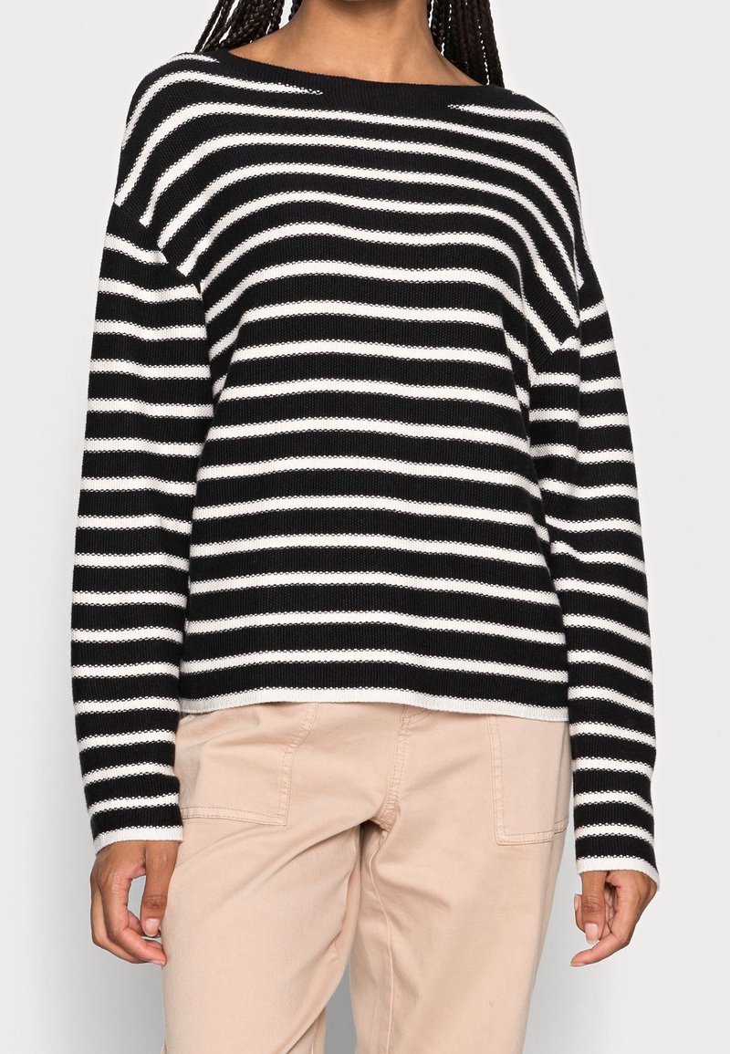 Schwarz-weiß gestreifter Pullover mit einem Bootsausschnitt und herunterfallenden Schultern, gestrickte Textur, kombiniert mit hellbeigen Hosen.