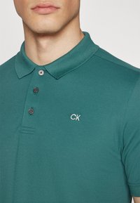Tirkizna polo majica izrađena od teksturiranog materijala, sa ovratnikom, tri crna dugmeta i izvezenim "CK" logom na lijevoj strani prsnog koša.