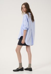 Světle modrá oversized košile s ohrnutými rukávy, spárovaná s černou sukni s jemným proužkem. Modelka má na sobě šedé vzorované ponožky a černé loafers.