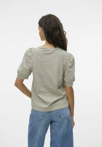 Γυναίκα με μακριά σκούρα μαλλιά, φορώντας μπλούζα με μπεζ και μαύρες ρίγες και φουσκωτά μανίκια, καθώς και μπλε τζιν, εμφανίζεται από πίσω σε λευκό φόντο.