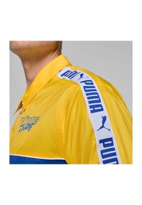 Żółta i niebieska sportowa kurtka z siateczkowej tekstury, z zapięciem na zamek i kontrastowymi paskami po bokach z logo "PUMA".