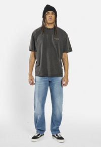 Σκούρο γκρι oversized T-shirt με διακριτικό κίτρινο λογότυπο, συνδυασμένο με ανοιχτό μπλε τζιν και μαύρα παπούτσια. Το μοντέλο φοράει ένα μαύρο σκουφί.