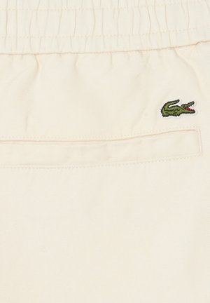 Pantalones de tela beige con cintura elástica, bolsillo trasero de ojal y logo de cocodrilo verde bordado cerca del bolsillo.