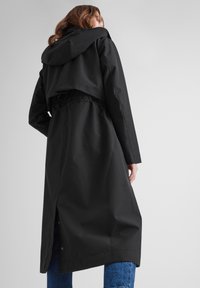 Black waterproof trench coat with a hood, cinched waist, and back vent detail. Smooth texture and long sleeves. Styled with blue denim.