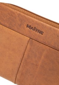 Brown leather wallet with textured surface, embossed "MAÎTRE" logo, and a zip closure. Features card slots on the front.