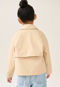 Lichtbeige cropped jack met een brede kraag en een gelaagd achterontwerp, gecombineerd met lichtblauwe denim jeans.