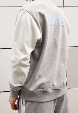 Person wearing a grey Nike jacket with white sleeves and matching grey sweatpants, standing against a tiled wall.