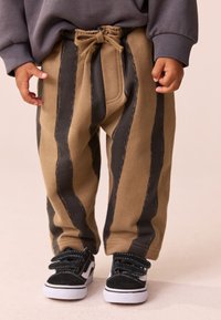 Beigefarbene Jogginghosen mit dunklen vertikalen Streifen, Tunnelzug an der Taille und Seitentaschen. Kombiniert mit schwarz-weißen Sneakers mit Klettverschlüssen.