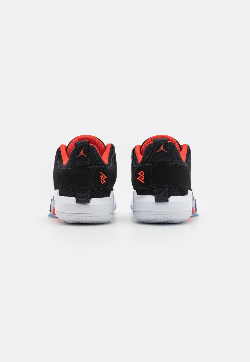Baskets en daim noir avec des accents rouges, semelles en caoutchouc blanche et détails texturés. Présente un onglet au talon arrière et une marque distinctive.