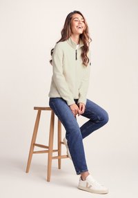 Light beige fleece jacket with a zip front and black accents, paired with blue jeans and white sneakers with beige details, sitting on a wooden stool.