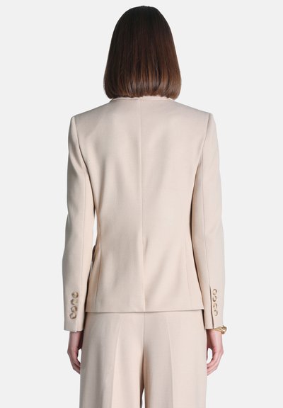 Femme aux cheveux bruns mi-longs portant un costume sur mesure beige clair, vue de dos sur un fond uni.