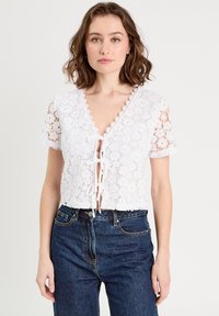 Haut court en dentelle blanche avec des manches courtes, des bords festonnés et un détail noué à l'avant. Associé à un jean en denim bleu foncé taille haute.