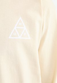 Beige bomull T-skjorte med et hvitt trekantet logo som har bokstavene H, U og F inni trekantene. Myk tekstur, rund halsdesign.