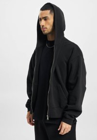 Sort zip-up hoodie lavet af blødt stof, med en stor hætte, frontlommer og afslappet pasform, vist på en model.