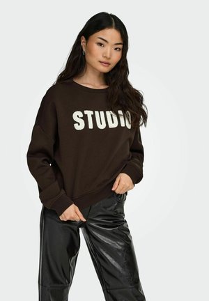 Sweatshirt marron avec le texte "STUDIO" en blanc, coupe décontractée, poignets côtelés, associé à un pantalon en cuir synthétique noir, texture lisse et finition brillante.