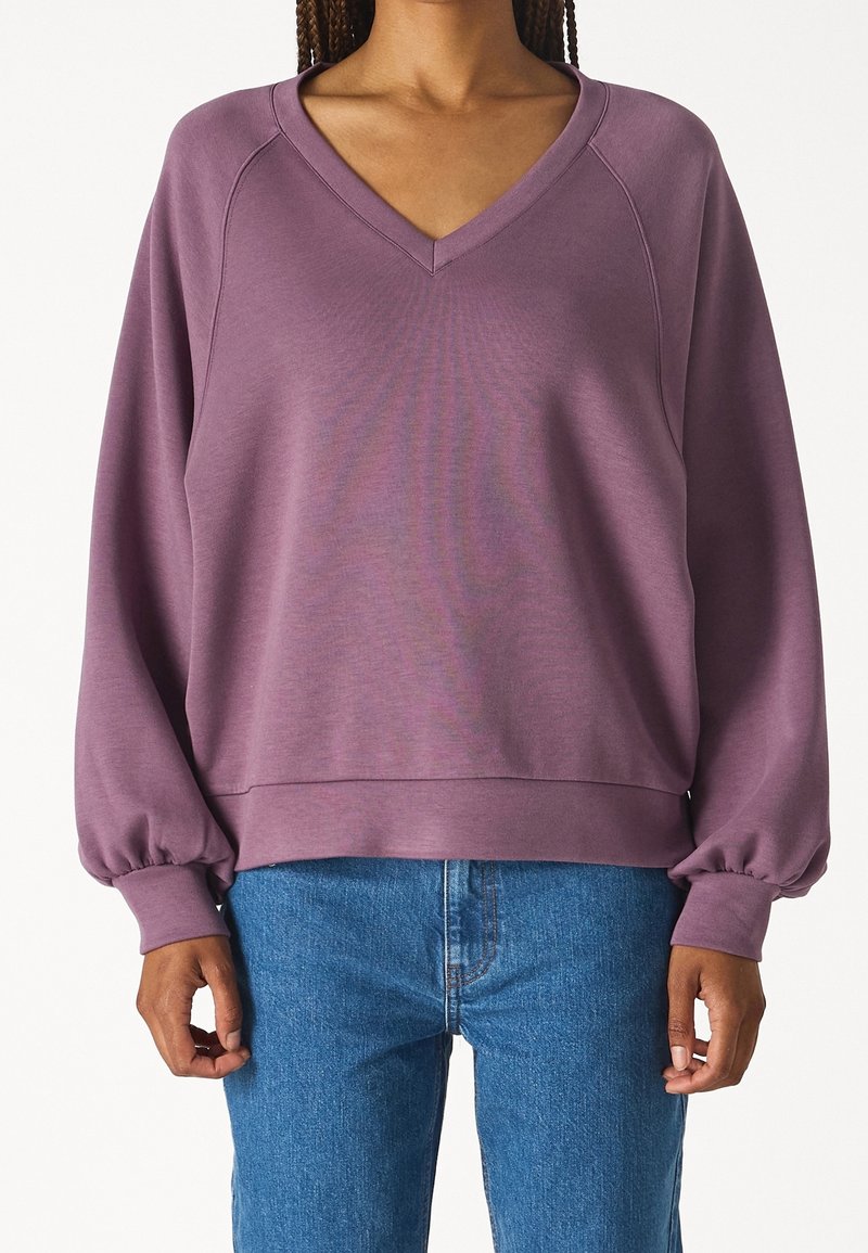 Personne portant un sweat ample mauve à col en V avec des manches ballon et un jean bleu, debout devant un fond blanc.