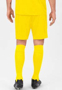Gelbe Fußballshorts und knielange Socken mit strukturiertem Muster, kombiniert mit schwarzen Fußballschuhen, von hinten betrachtet.
