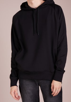Person trägt einen schlichten schwarzen Hoodie mit Kordeln und dunkelgraue Jeans, schaut nach vorne vor neutralem Hintergrund.