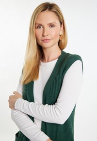 Zelena pletena prsluk preko bijele majice dugih rukava. Prsluk ima teksturirani rebrasti dizajn i otvoreni prednji dio. Kosa je ravna i plava.