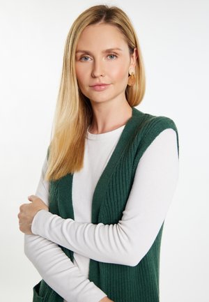 Zielona dzianinowa kamizelka na białej bluzce z długim rękawem. Kamizelka ma fakturowany, prążkowany wzór i otwarty przód. Włosy są proste i blond.