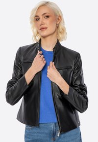 Schwarze Lederjacke mit Stehkragen, Reißverschluss vorne und flachen Brusttaschen. Trägt sie über einem blauen Pullover, kombiniert mit Jeans. Glatte Textur.